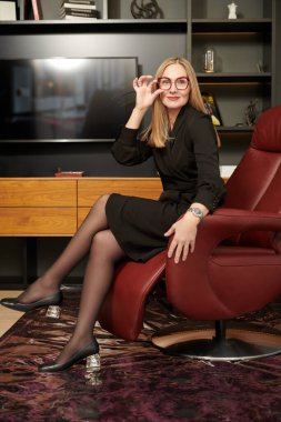 Modern bir ofiste pahalı bir deri koltukta oturan ve gülümseyen şık, orta yaşlı güzel bir kadının fotoğrafı. Zarif siyah elbise ve gözlüklü başarılı bir iş kadını..