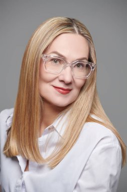 Klasik beyaz bluz ve gözlük takmış orta yaşlı mutlu bir iş kadınının portresi. Gri stüdyo geçmişi. Gözlük stili. 