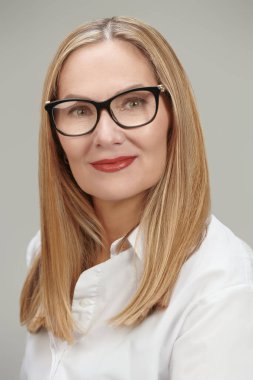 Klasik beyaz bluz ve gözlüklü, orta yaşlı, mutlu bir iş kadınının fotoğrafı. Gri stüdyo geçmişi. Gözlük stili. 