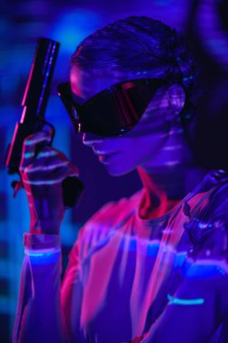 Fütürist aynalı gözlüklü ve tulumlu mükemmel siber punk kızı kırmızı ışıkta duruyor ve elinde silahla ateş yansıtıyor. Geleceğin siber dünyası. Sanal oyunlar.