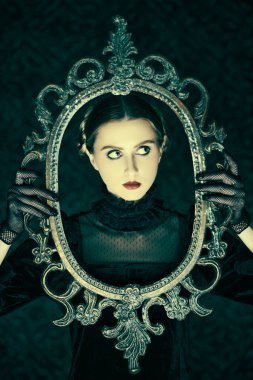 Zarif siyah kadife bir elbise ve dantel eldivenli dramatik esmer bir kızın portresi, gecenin karanlığında elinde ayna çerçevesi olan eski bir arka planda duruyor. Gotik roman, mistisizm..