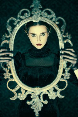 Zarif siyah kadife bir elbise ve dantel eldivenli dramatik esmer bir kızın portresi, gecenin karanlığında elinde ayna çerçevesi olan eski bir arka planda duruyor. Gotik roman, mistisizm..