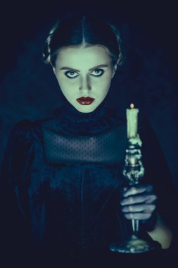 Portrait of a dramatic brunette girl in an elegant black velvet dress and lace gloves, who stands in the night on a dark vintage background with a burning candle in her hands. Gothic novel, mysticism. clipart