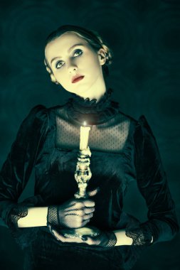 Zarif siyah kadife elbise ve dantel eldivenli dramatik esmer bir kızın portresi, gecenin karanlığında elinde yanan bir mumla eski bir arka planda duruyor. Gotik roman, mistisizm..
