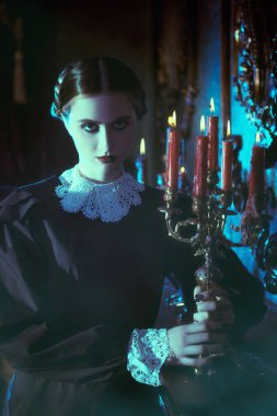 Zarif siyah kadife elbiseli dramatik esmer bir kızın portresi, gecenin karanlığında elinde mumlarla eskimiş bir arka planda duruyor. Gotik roman, mistisizm..