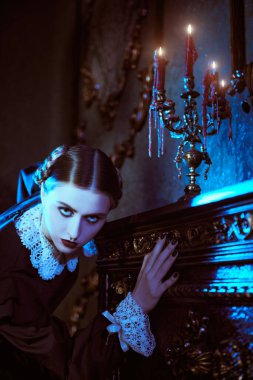 Gothic novel. A gloomy brunette girl in a strict black dress with a white collar stands in a threatening pose by the fireplace in a gloomy mansion at night. The Addams Family. clipart