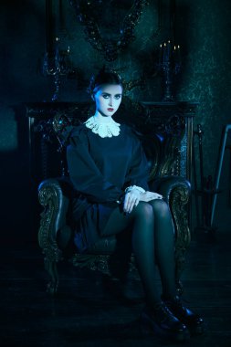 Kasvetli bir şatoda, koltukta oturan, katı siyah elbiseli, beyaz yakalı, tuhaf, esmer bir kızın tam boy portresi. Mistik gotik roman, korku. Addams Aile Stili. 