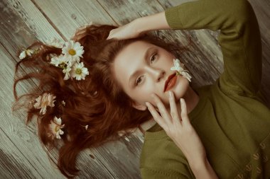 Bahar ve yaz havası. Dalgalı kızıl saçlı güzel bir kız saçlarında beyaz çiçeklerle zeytin yeşili bir elbiseyle yerde poz veriyor. Uzayı kopyala Kadın güzelliği ve moda.