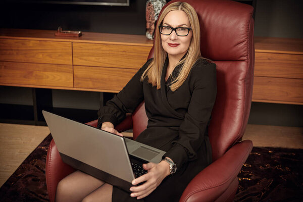 Красивая стильная женщина средних лет сидит в дорогом кожаном кресле в современном офисе и улыбается. Успешная деловая женщина в элегантном черном платье и очках.