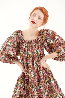 Çiçekli bir elbisenin içinde beyaz bir duvara karşı duran narin makyajlı güzel bir kızıl kızın portresi. Bahar ve yaz modası. Romantik kadınsı stil.