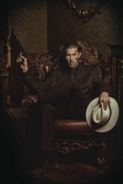 Zarif siyah takım elbiseli ve beyaz şapkalı sert bir gangster lüks bir dairede elinde silahla bir sandalyede oturuyor. Retro tarzı. Mafyanın suç dünyasına. Dedektif filmi.