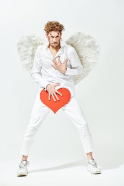 Çekici Aşk Tanrısı, elini kalbine yakın tutarak baştan çıkarıcı bir şekilde kameraya bakar. Beyaz stüdyo arka planında tam boy portre. Aşk konsepti. Fantezi. Sevgililer Günü.