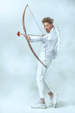 Sevgililer Günü. Cesur aşk tanrısı Eros 'un tam boy portresi. Yüzünde yaralar var. Kurbanına ok atma niyetinde. Fantezi. Stüdyo beyaz arka plan. Aşk konsepti.