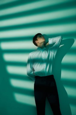 Erkek Moda ve Güzellik. Beyaz gömlekli yakışıklı bir gencin sanat portresi ve beyaz ve akuamarin renkli stüdyo ışıklarıyla poz veren siyah pantolon. Işık ve gölge. Boşluğu kopyala.