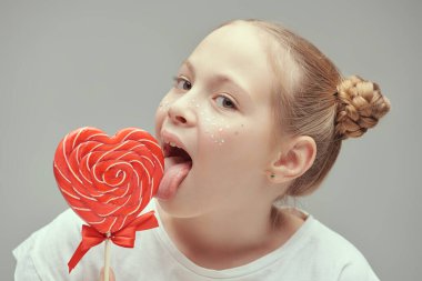 Şirin çilli komik mutlu kız büyük kalp şeklinde bir lolipopu yalıyor. Çocuklar, mutlu çocukluk, çocukların duyguları. Gri stüdyo geçmişi.