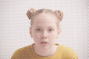 A surprised girl wearing casual olive pullover stands behind the lattice. Emotions and psychology of kids. clipart