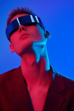 Moda konsepti. Mis mavisi ve kıpkırmızı ışıkta aynalı fütürist gözlüklerle poz veren şık bir gencin portresi. Mavi stüdyo geçmişi. Gözlük stili. 