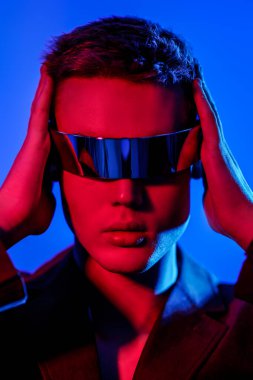 Mis mavisi ve kıpkırmızı ışıkta aynalı fütürist gözlüklerle poz veren şık bir gencin portresi. Mavi stüdyo geçmişi. Gözlük stili. Moda konsepti. 