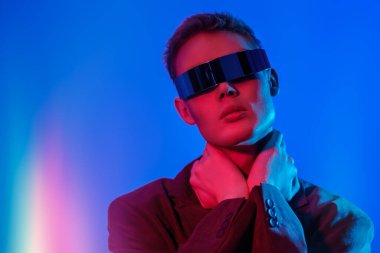 Moda konsepti. Mis mavisi ve kıpkırmızı ışıkta aynalı fütürist gözlüklerle poz veren şık bir gencin portresi. Fotokopi alanı olan mavi stüdyo. Gözlük stili. 