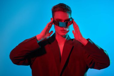 Modaya uygun bir modelin, aynalı mavi ve kırmızı ışıkta fütürist gözlüklerle poz vermesi. Mavi stüdyo geçmişi. Gözlük stili. Yüksek Moda.