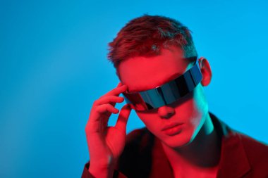 Mis mavisi ve kıpkırmızı ışıkta aynalı fütürist gözlüklerle poz veren şık bir gencin portresi. Fotokopi alanı olan mavi stüdyo. Gözlük stili. Moda konsepti. 