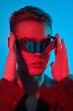 Mis mavisi ve kıpkırmızı bir ışıkta aynalı fütürist gözlüklerle poz veren şık bir gencin fotoğrafı. Mavi stüdyo geçmişi. Gözlük stili. Moda konsepti. 