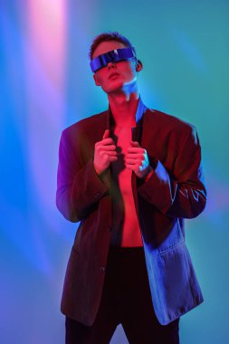 Moda konsepti. Mis mavisi ve kıpkırmızı ışıkta aynalı fütürist gözlüklerle poz veren şık bir gencin portresi. Mavi stüdyo geçmişi. Gözlük stili. 