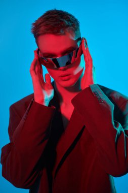 Moda konsepti. Mis mavisi ve kıpkırmızı ışıkta aynalı fütürist gözlüklerle poz veren şık bir gencin portresi. Mavi stüdyo geçmişi. Gözlük stili. 
