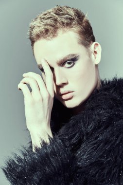 Kısa saç kesimi ve siyah dumanlı gözleriyle stüdyoda siyah kürklü poz veren şık bir gencin portresi. Moda ve erkek güzelliği. Makyaj ve saç stili, kozmetik..