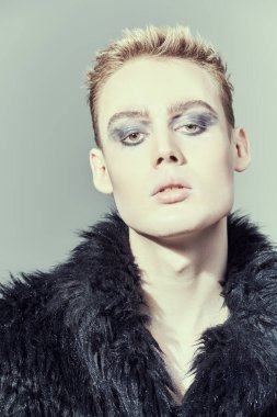 Makyaj, saç stili, makyaj malzemeleri. Kısa saç kesimi ve siyah dumanlı gözleriyle stüdyoda siyah kürklü poz veren şık bir gencin portresi. Moda ve erkek güzelliği. 