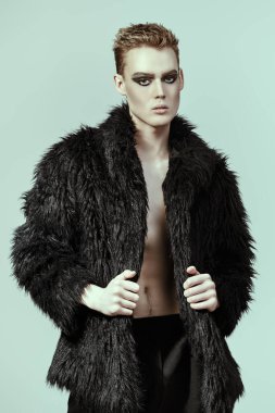 Kısa saç kesimi ve siyah dumanlı gözleriyle stüdyoda siyah kürklü poz veren şık bir gencin fotoğrafı. Makyaj, saç stili, makyaj malzemeleri. Moda ve erkek güzelliği. 