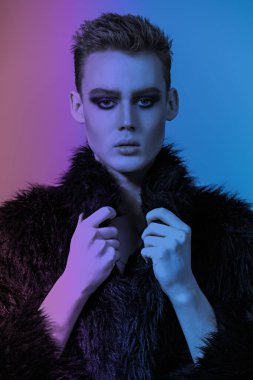 Kısa saç kesimi ve siyah dumanlı gözleriyle stüdyoda siyah kürklü poz veren şık bir gencin portresi. Moda ve erkek güzelliği. Makyaj, saç stili, makyaj malzemeleri. Karışık renkler, sanat.