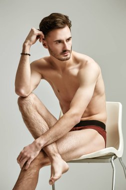 Sandalyenin üzerinde beyaz bir stüdyo arka planına karşı erkek donu içinde çıplak poz veren yakışıklı bir sporcunun portresi. İç çamaşırı ve erkek güzelliği..