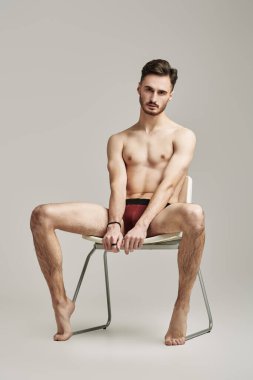 Sandalyenin üzerinde beyaz bir stüdyo arka planına karşı erkek donu içinde çıplak poz veren yakışıklı bir atletin tam boy portresi. İç çamaşırı ve erkek güzelliği..