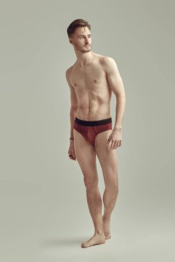 Beyaz arka planda erkek donu giymiş çıplak poz veren yakışıklı bir atletin uzun bir stüdyo portresi. İç çamaşırı ve erkek güzelliği..