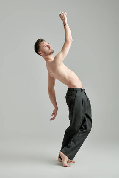 Полнометражный портрет красивого атлетичного брюнетки с голым туловищем, танцующим на сером фоне студии. Мужское здоровье и красота.