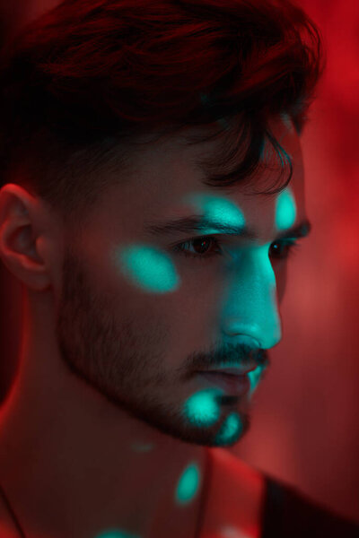 An art portrait of a masculine handsome brunette man standing in a dark room with red lighting. Men's emotions and beauty. Beams of light fall on him.