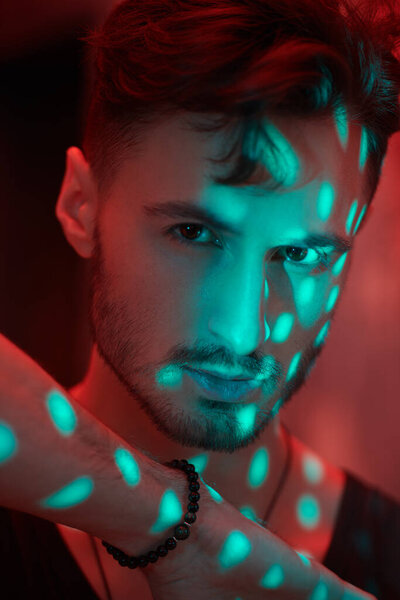 Men's emotions and beauty. Art portrait of a masculine handsome brunette man, who is looking ahead warily and confidently, standing in a dark room with red lighting. Beams of light fall on him.