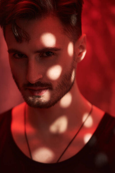 Men's emotions and beauty. Art portrait of a masculine handsome brunette man, who is looking ahead warily and confidently, standing in a dark room with red lighting. Beams of light fall on him.
