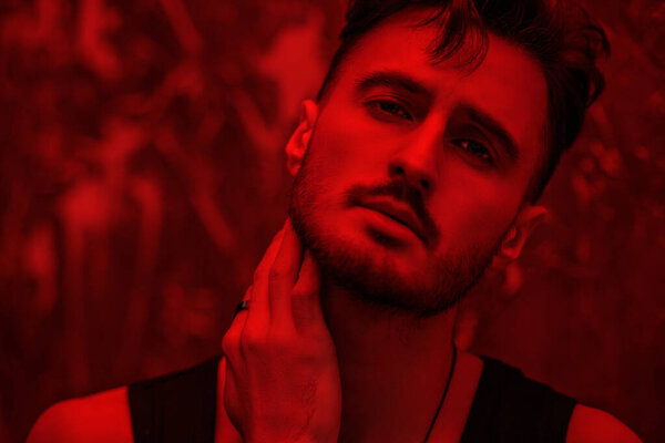 Men's emotions and beauty. Art portrait of a masculine handsome brunette man, who is looking ahead warily and confidently, standing in a dark room with red lighting. Beams of light fall on him.