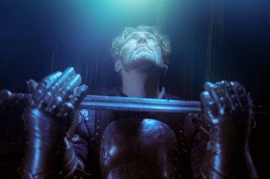 Zırhlı yakışıklı bir ortaçağ şövalyesi kalede yükseltilmiş bir kılıçla duruyor ve yukarı bakıyor. Şövalye romantizminden bir sahne. Tarihi yeniden yapılandırma.