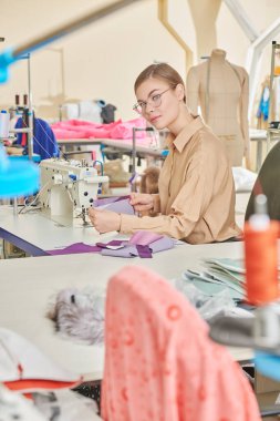 Moda endüstrisi. Mutlu yaratıcı genç bir terzi kız atölyede yeni bir ürün dikiyor. Giysi imalatı.