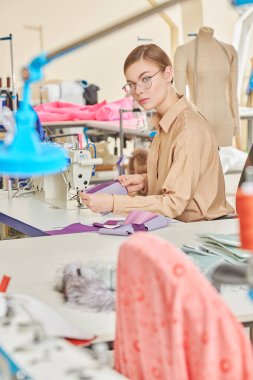 Moda endüstrisi. Yaratıcı genç bir kızın mutlu bir fotoğrafı atölyede yeni bir ürün dikiyor. Giysi imalatı.