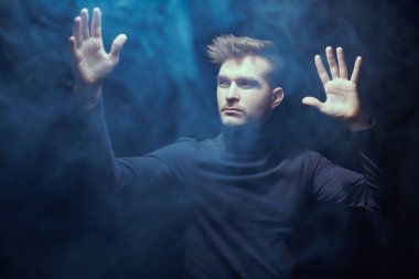 Sakin ve odaklanmış bir adam sisle çevrili siyah bir arka planda durur ellerini kaldırır ve yüzünde derin bir ifadeyle ışıltıya bakar. Psikoloji, telepati..