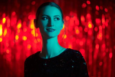 Gece makyajı ve parlak pullu gece elbisesiyle kırmızı folyo perdelere poz veren güzel bir kadının portresi. Şenlikli bir akşam stili. Noel ve Yeni Yıl.