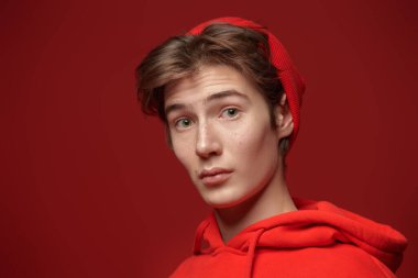 Kırmızı eşofmanlı ve kırmızı şapkalı yakışıklı bir çocuk kameraya bakıyor. Kırmızı arkaplanda fotokopi alanı olan bir stüdyo resmi. Gençler, duygular.