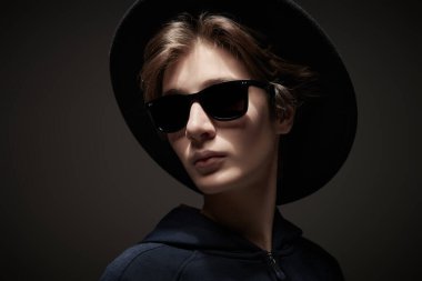 Siyah kapüşonlu, şapkalı ve güneş gözlüklü havalı bir gencin fotoğrafı. Karanlık arka plan. Modern gençler, yaşam tarzı..