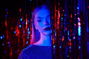 A trendy blonde girl with shiny glitter makeup and a sparkling evening dress stands among red foil curtains. Night club, disco, neon lighting. Glitter, fashion and style. 