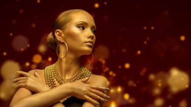 Noel güzeli kız. Altın tenli, makyajlı, altın takılar takmış altın sarısı parlak ışıklarla süslü kırmızı bir arka planda poz veren güzel bir genç kadın. Altın lüks konsept. Boşluğu kopyala.