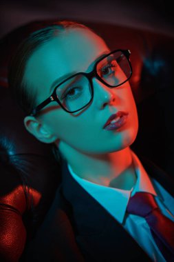Takım elbiseli, kravatlı ve deri koltukta gözlüklü şık, kendine güvenen bir kadının portresi. Karışık ışıklandırma. Lüks bir iş tarzı. Gözlük modası.